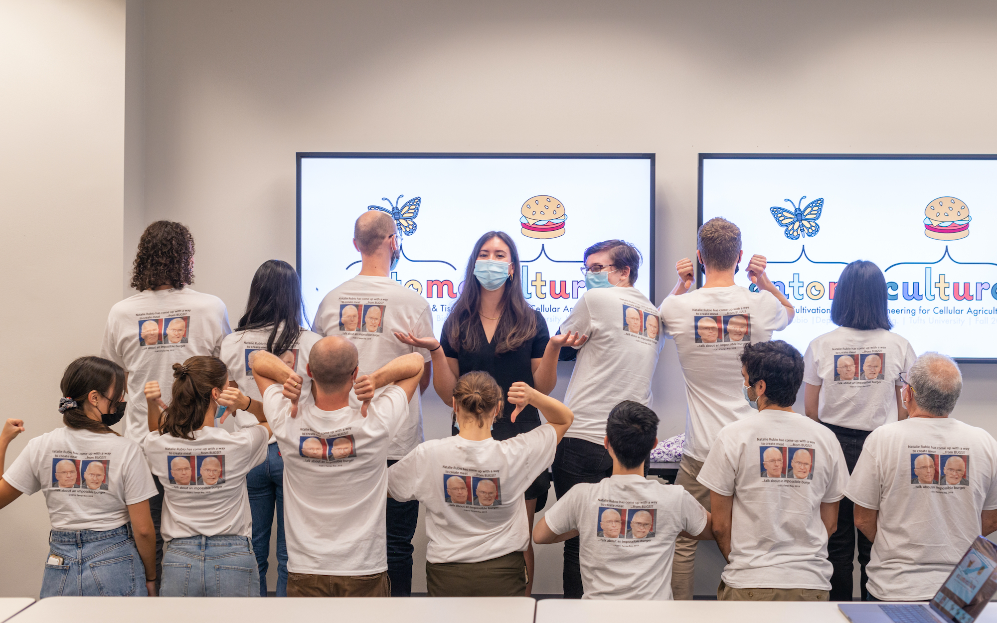 group of 14 photo of everyone (except for main researcher in center) wearing the same white t-shirt turned around pointing to the back of the shirt that has a meme